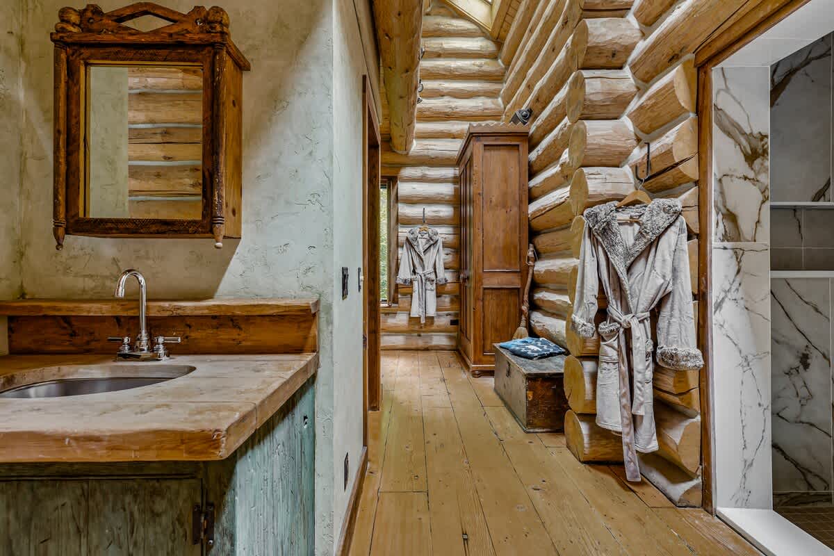 Rustic bathroom vanity area with log cabin walls and wooden floors, featuring a custom wooden countertop with an integrated sink and a decorative mirror, leading into a tiled shower.