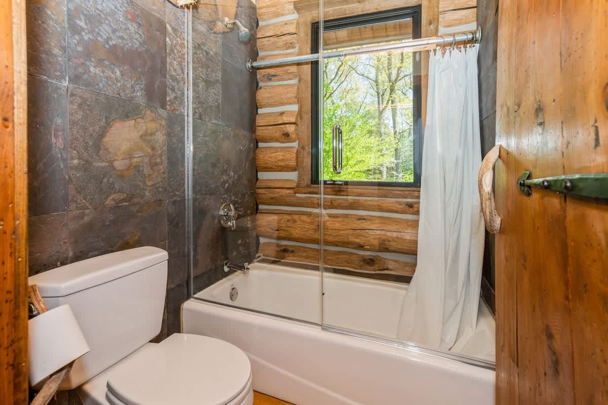 Rustic bathroom with dark slate tiled shower walls and a white bathtub, featuring a toilet and a window offering a view of the surrounding forest.