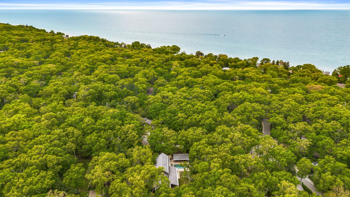 A wide aerial shot reveals the expansive property, densely forested with vibrant green trees, situated near a large, calm body of water under a clear blue sky, highlighting the secluded natural setting.