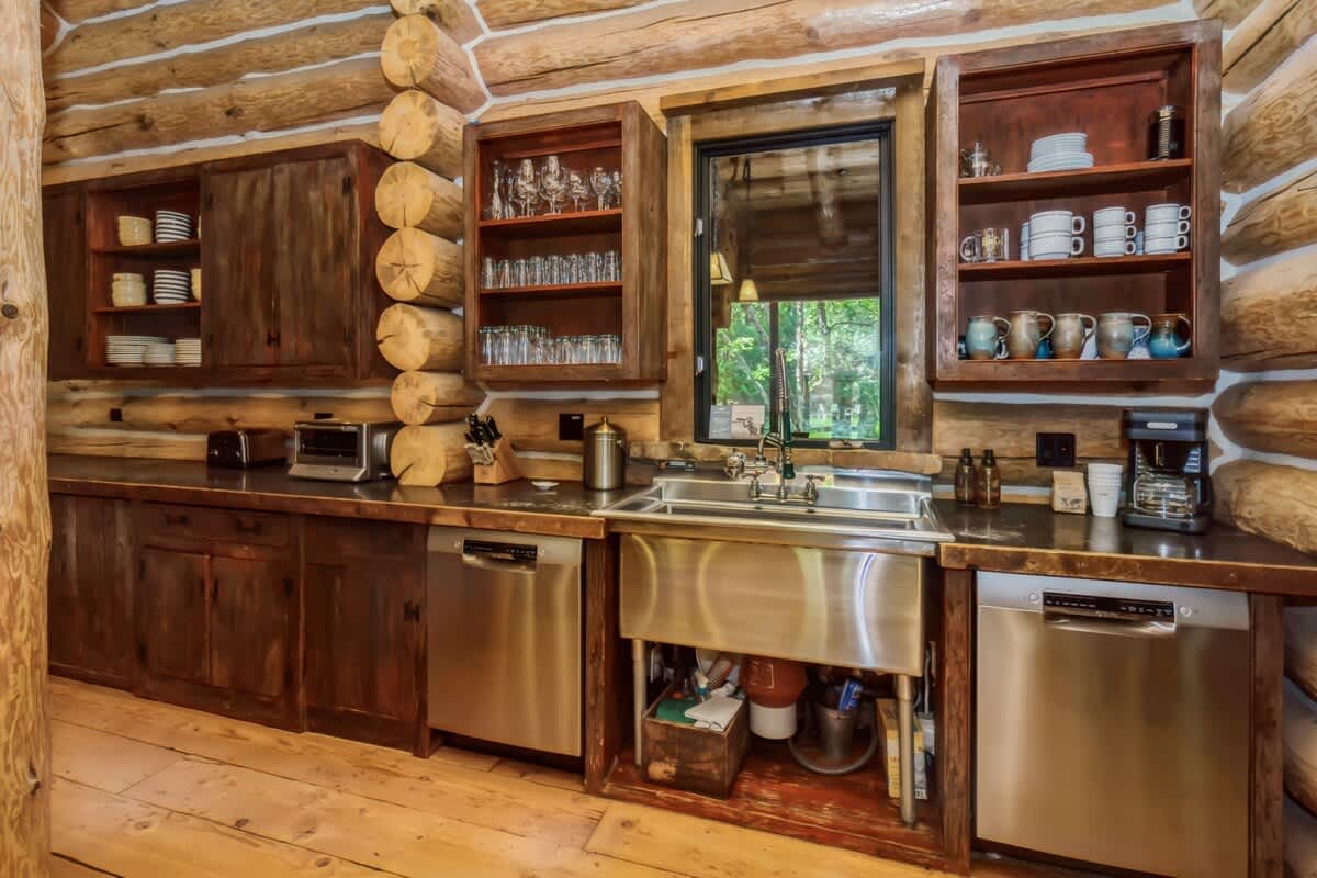 A detailed view of the log cabin kitchen's wet bar area, featuring a large stainless steel sink, two dishwashers, dark wooden countertops, and open shelving with various dishware.