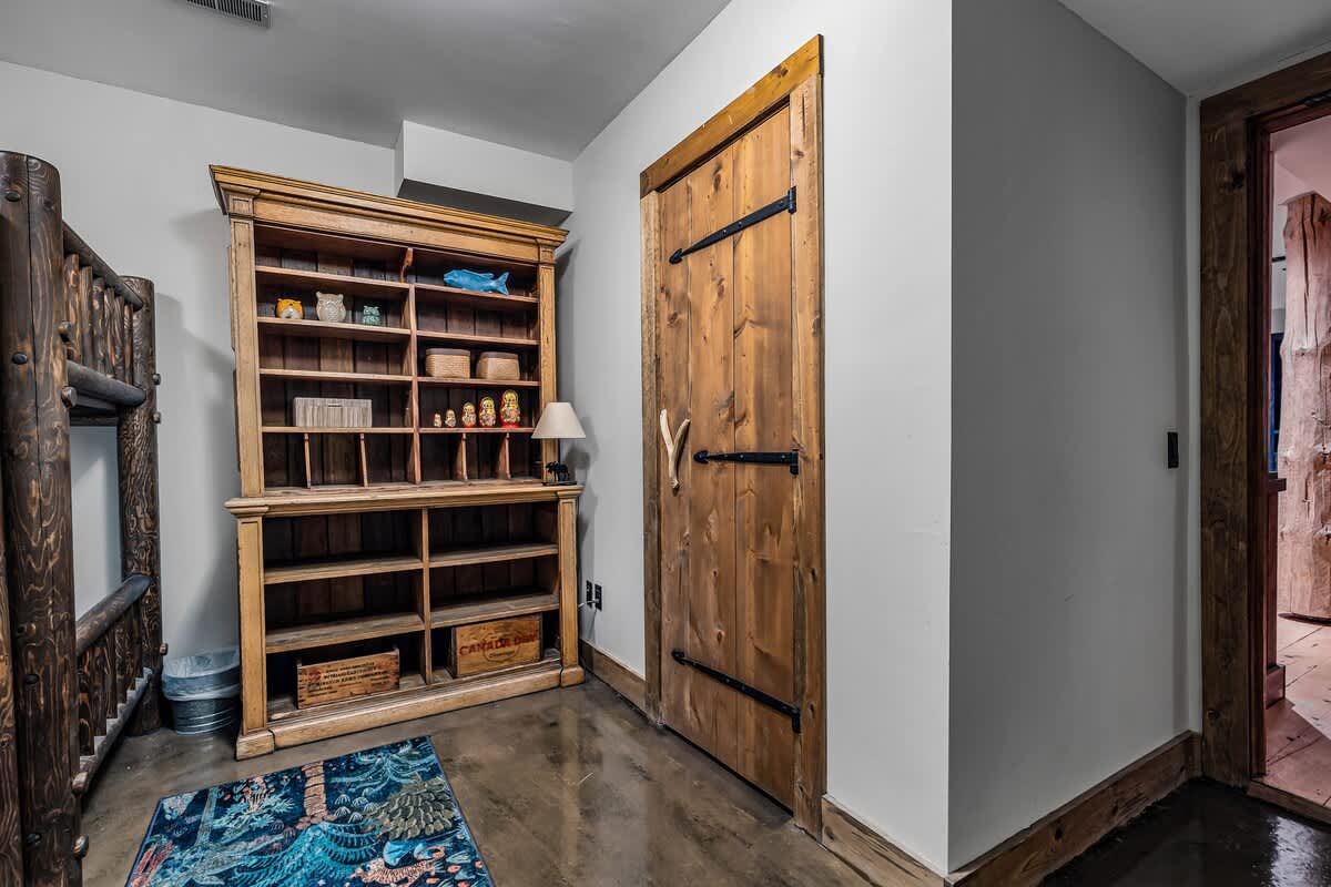 Rustic hallway or entrance area featuring a tall wooden bookshelf filled with decorative items, a sturdy wooden door with black hardware, and a colorful patterned rug on a concrete floor.