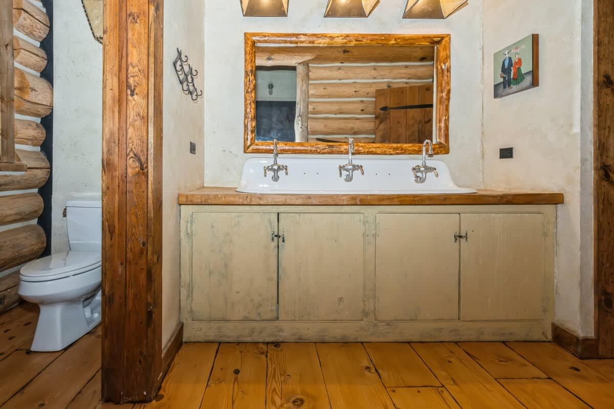 Rustic bathroom showcasing a wooden vanity with a trough sink and log-framed mirror, a toilet partially visible behind a wooden column, and warm hardwood floors, emphasizing the natural wood elements.