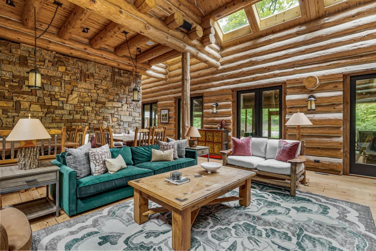 A spacious log cabin living room with exposed log walls and ceiling, featuring a dark green velvet sofa and a lighter sofa, arranged around a large wooden coffee table. A stone accent wall and a dining area are visible in the background, creating an open-concept feel.