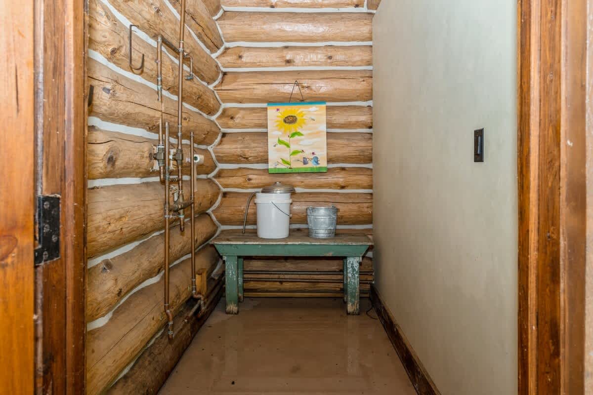 A rustic outdoor utility area features exposed log walls and visible copper plumbing, with a small green bench holding metal buckets. A colorful sunflower painting adds a touch of charm to the natural wood setting.