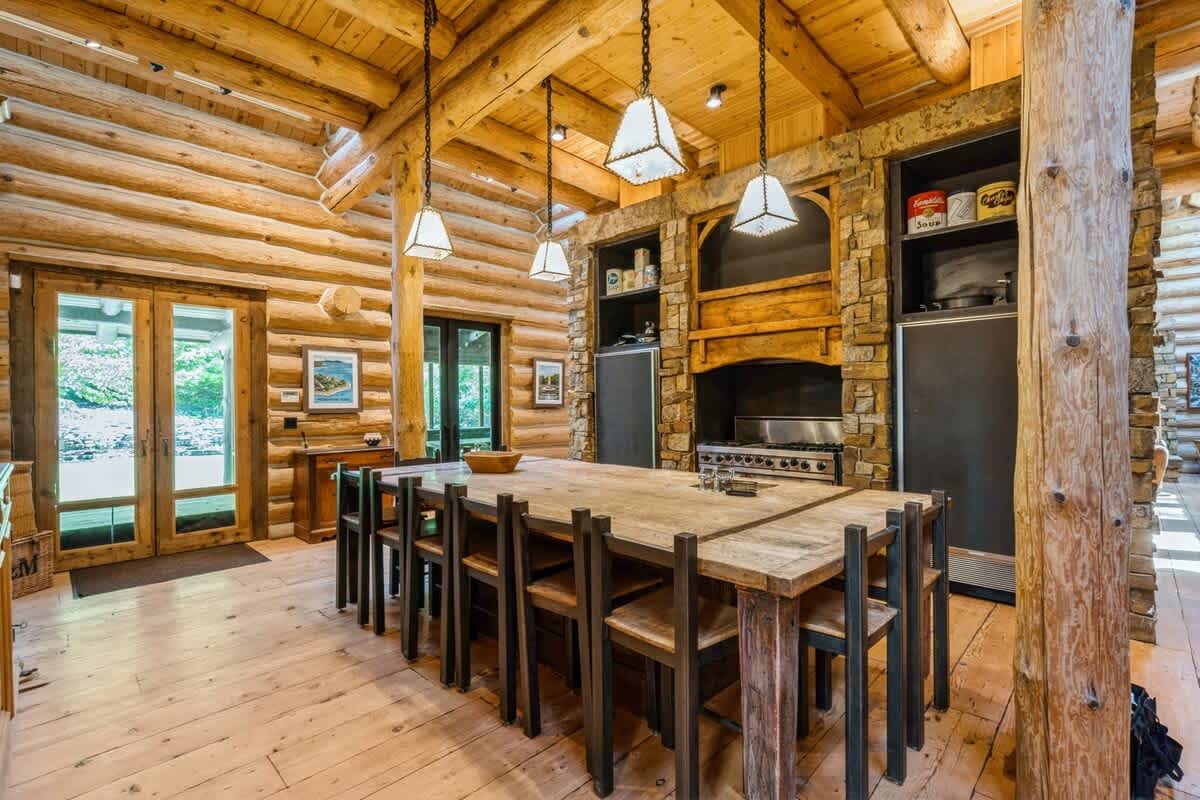 A rustic log cabin kitchen featuring a large wooden island that doubles as a dining table, surrounded by multiple chairs. The space boasts exposed log walls and ceiling, stone accents around the stove, and stylish pendant lighting.
