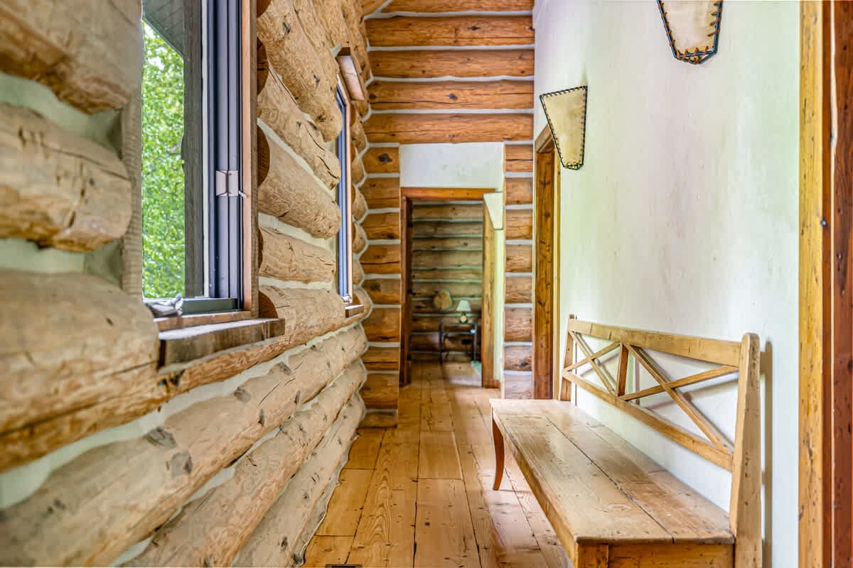 A rustic hallway with exposed log walls and light wooden floors, featuring a long wooden bench and leading to other rooms through open doorways.