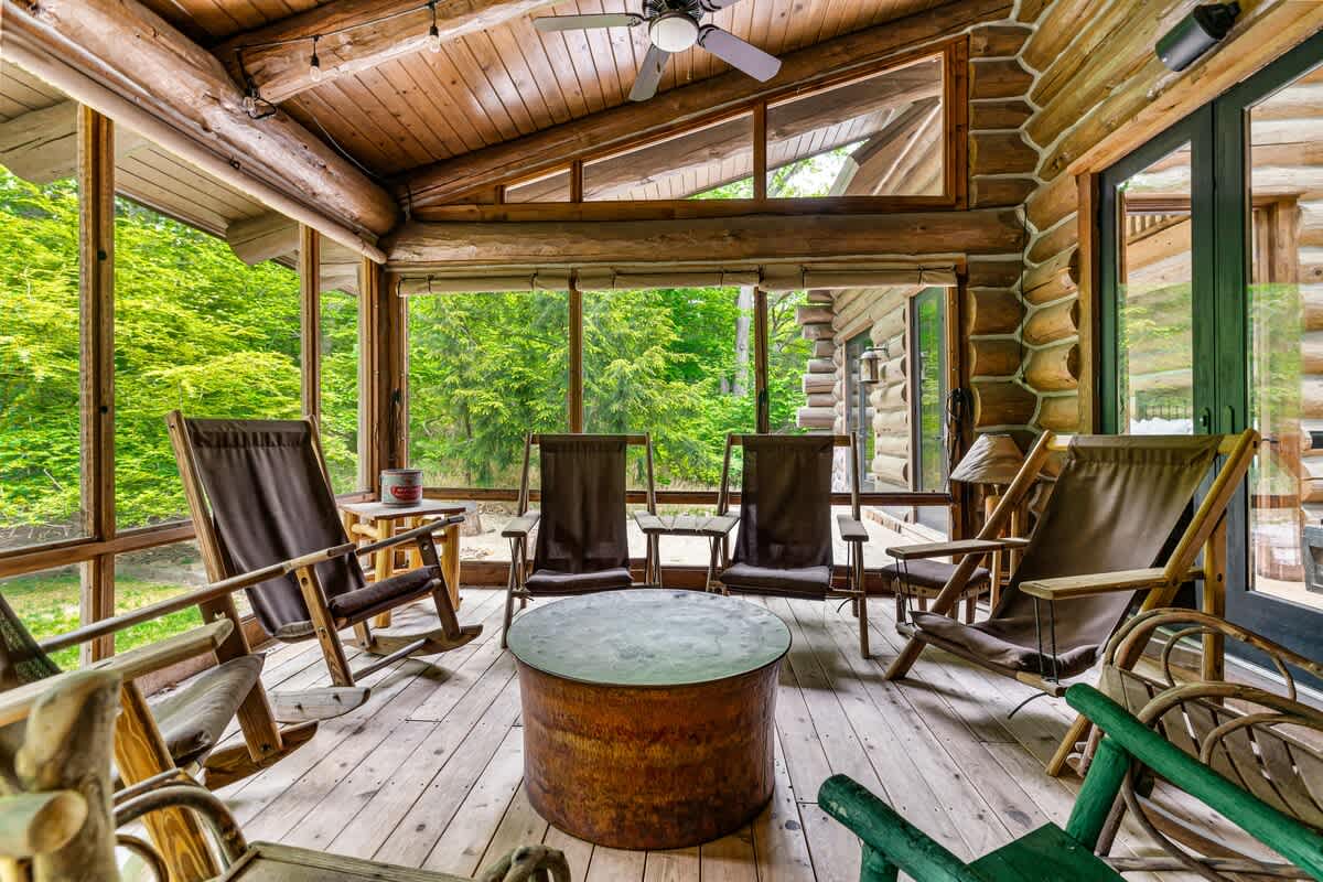 Inviting screened-in porch with log cabin walls and wooden floors, furnished with several comfortable rocking chairs and a round coffee table, perfect for relaxing amidst nature.