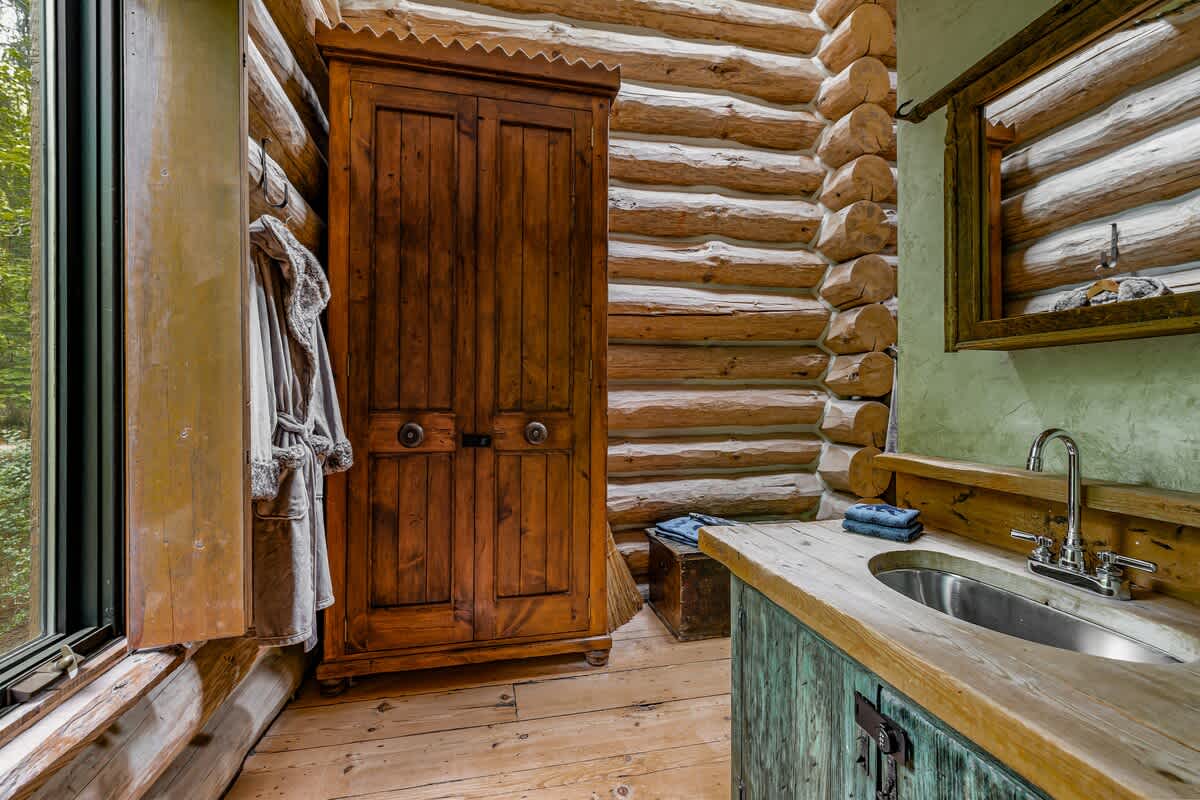Rustic bathroom featuring log walls, a wooden vanity with a metal sink, and a tall wooden cabinet, with a window offering a view of lush greenery.