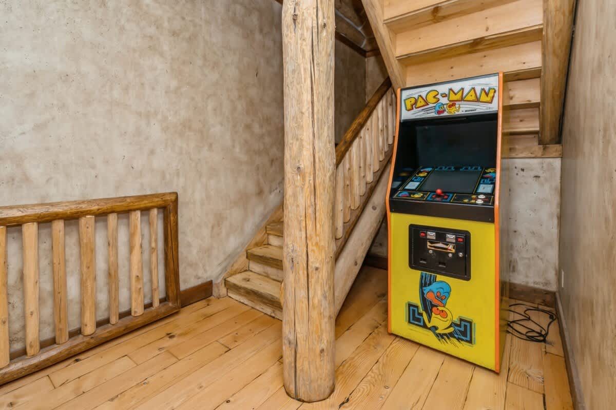 A classic yellow Pac-Man arcade machine stands beside a rustic wooden staircase with log newel posts and railings. The walls have a textured, light-colored finish, complementing the natural wood elements.