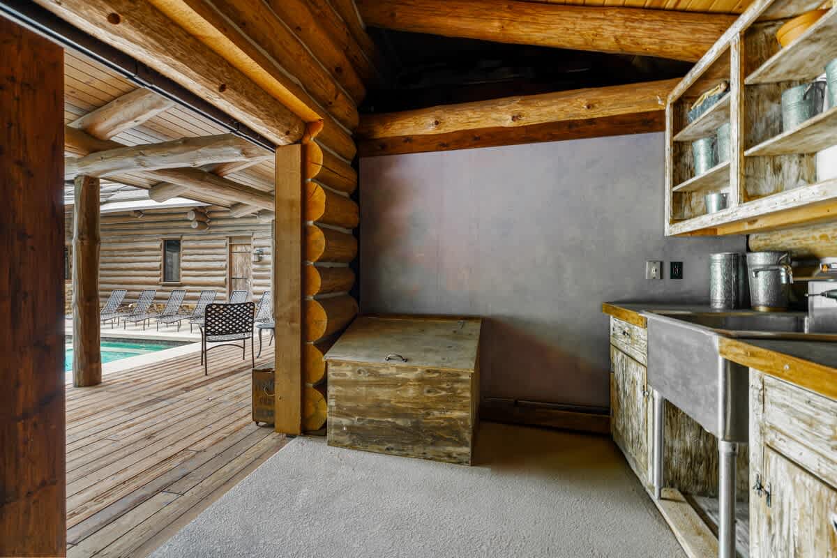 A rustic outdoor kitchen area, part of a log cabin structure, features a metal sink set into a wooden countertop with open shelving above. The space opens to a wooden deck overlooking the swimming pool, perfect for outdoor entertaining.