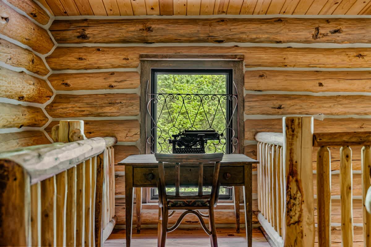 Charming loft workspace in a log cabin, featuring a small wooden desk with a vintage typewriter, surrounded by rustic wooden railings and a window overlooking the trees.