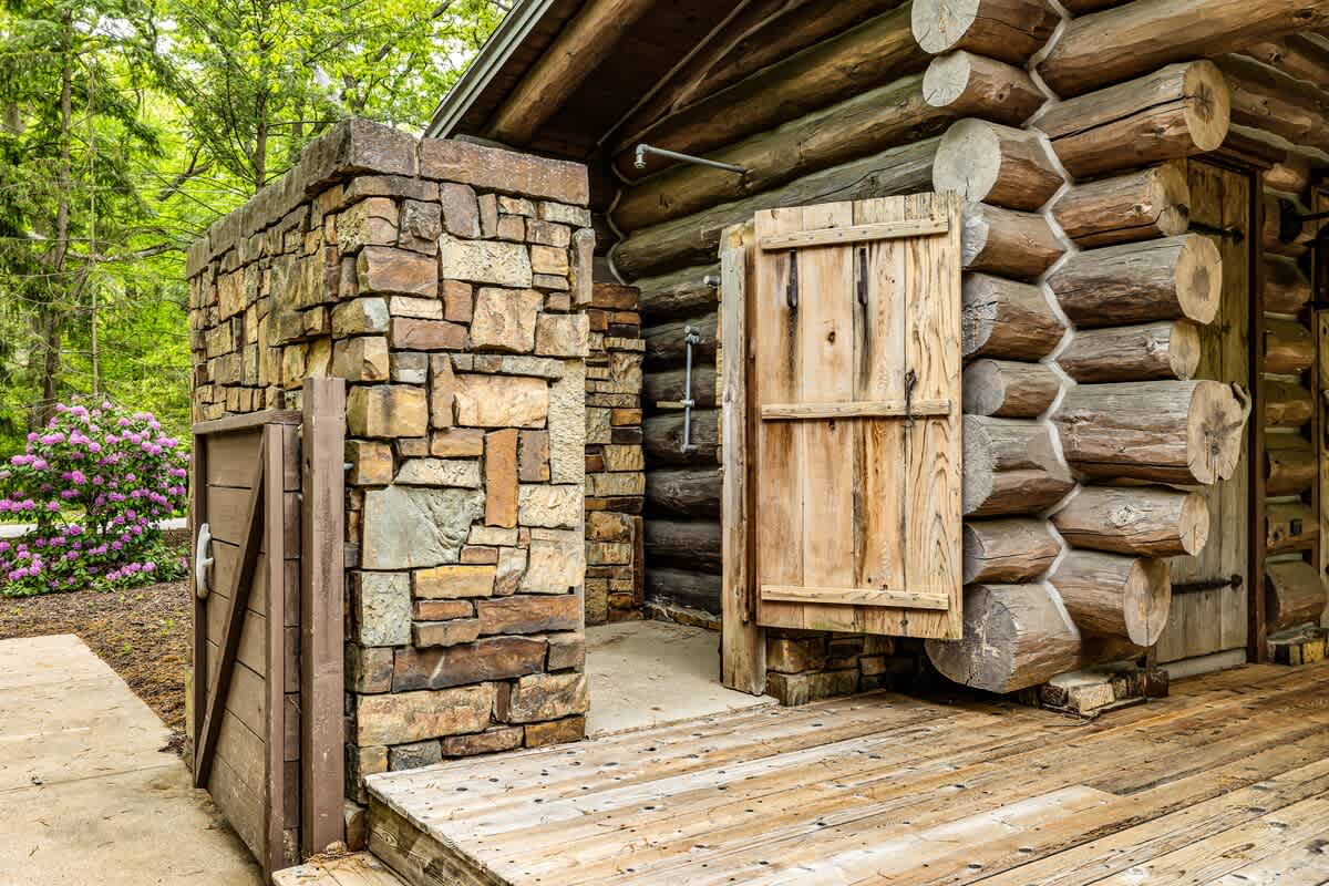 A rustic outdoor shower area with a wooden door and stone wall enclosure, adjacent to a log cabin with stacked timber construction. A wooden deck extends from the cabin.