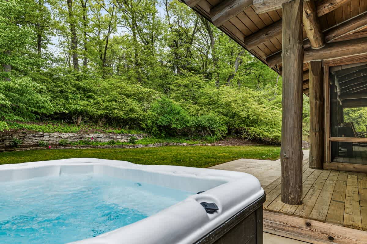A relaxing outdoor hot tub with bubbling water, situated on a concrete patio adjacent to a wooden deck and the log cabin exterior. The area is surrounded by a vibrant green lawn and dense forest, offering privacy and natural beauty.