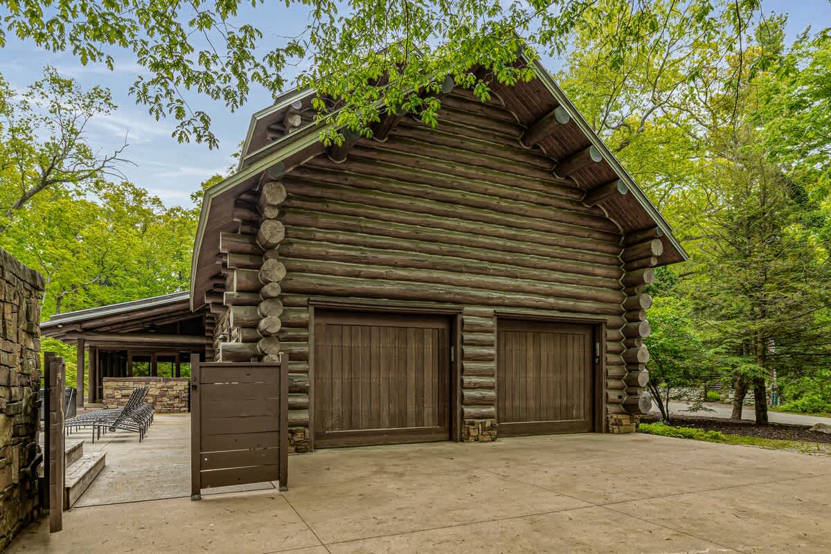 The exterior of a large log cabin garage featuring two rustic wooden garage doors and stone accents at the base, surrounded by lush green trees under a bright sky.