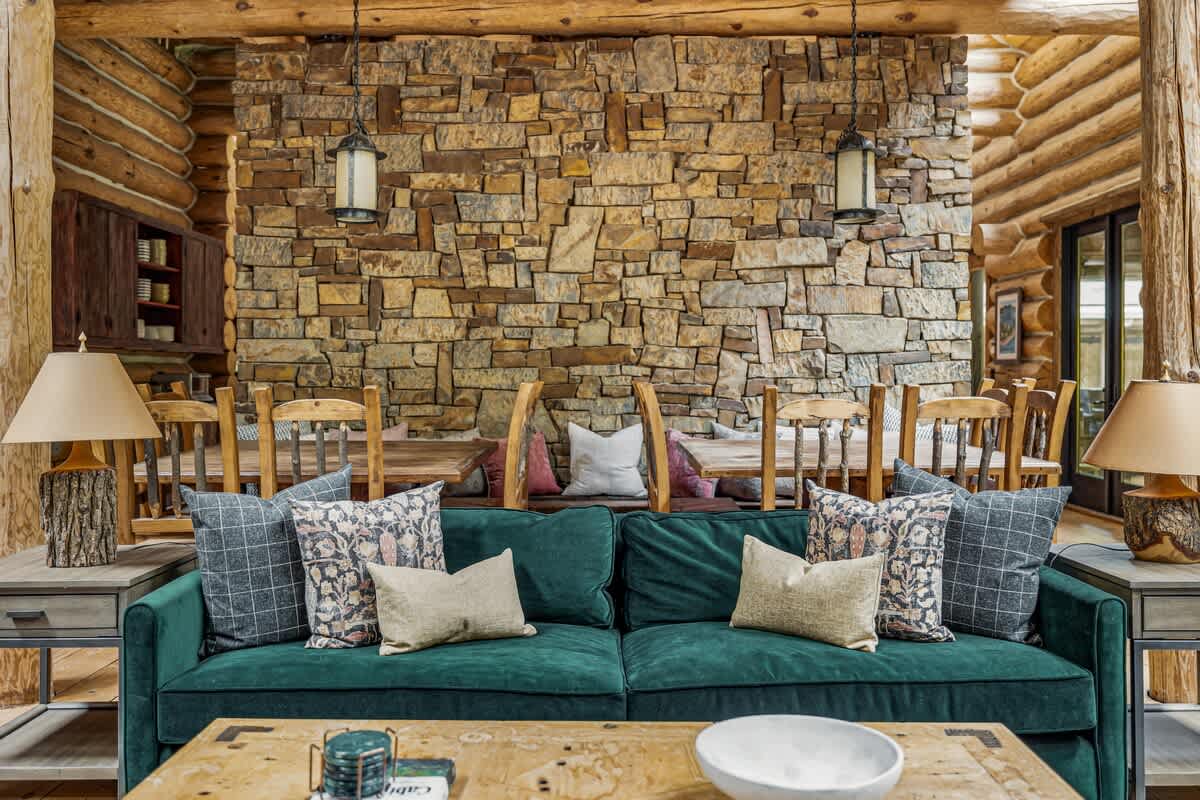 A close-up view of the living room, highlighting a dark green velvet sofa adorned with decorative pillows and a rustic wooden coffee table. A prominent stone accent wall and a dining area with wooden chairs are visible in the background, showcasing the cabin's cozy elegance.