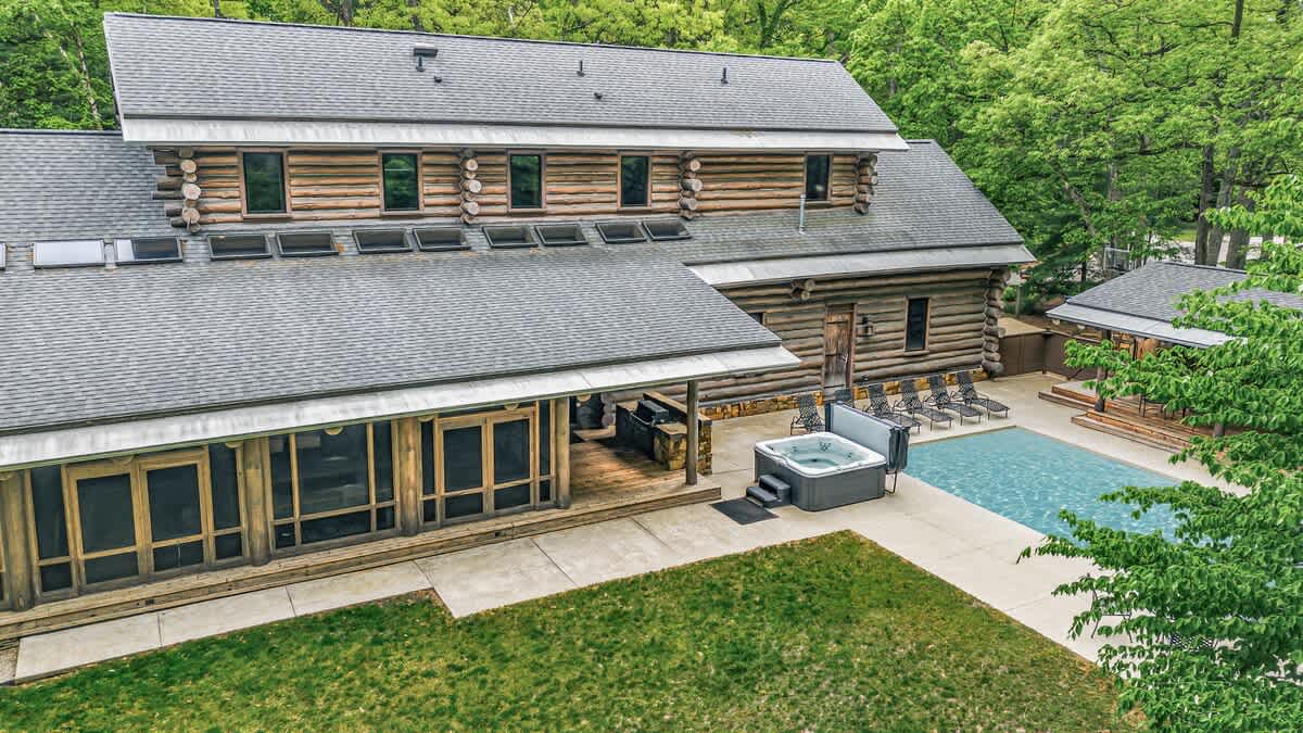 An aerial view showcases a sprawling log cabin property with a large main house and a separate pool house, both featuring gray shingle roofs. A sparkling outdoor pool and hot tub are visible on the concrete patio, surrounded by lush green trees.