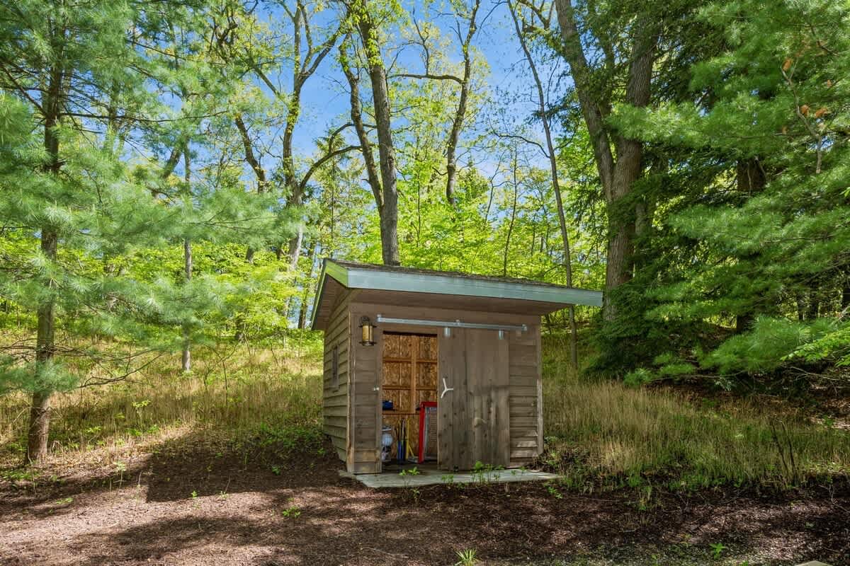 A small, rustic wooden shed with a sliding barn door and a single exterior light fixture, nestled on a grassy slope surrounded by tall trees and lush greenery.