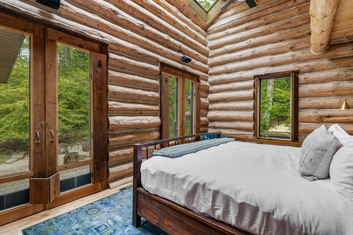 Bright log cabin bedroom with a wooden bed, white bedding, and a blue patterned rug, featuring large glass double doors and multiple windows that flood the room with natural light and offer forest views.
