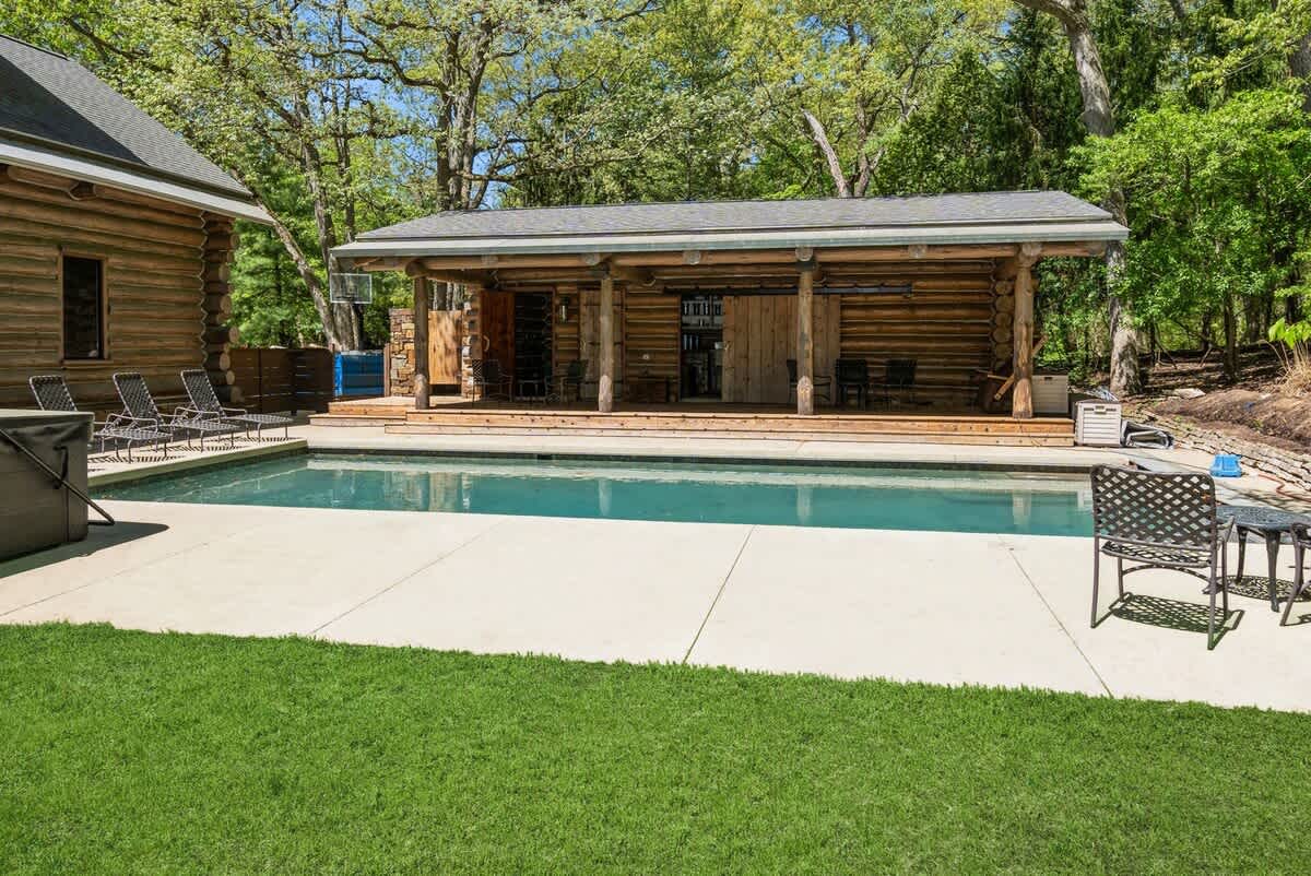 An inviting outdoor swimming pool is surrounded by a concrete patio and lush green lawn, with a rustic log cabin pool house in the background. Several lounge chairs are arranged for sunbathing, set against a backdrop of mature trees.