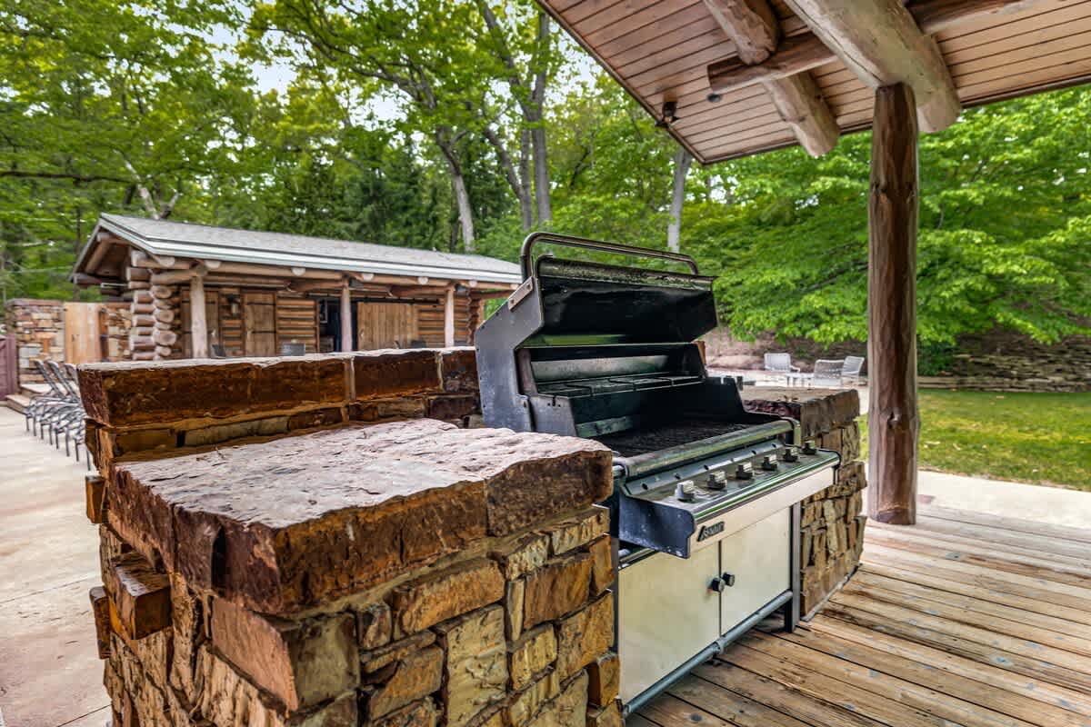 A robust outdoor grilling station built with natural stone, featuring a large gas grill under a covered wooden patio. Lush green trees and another log cabin structure are visible in the background, creating a perfect outdoor cooking space.