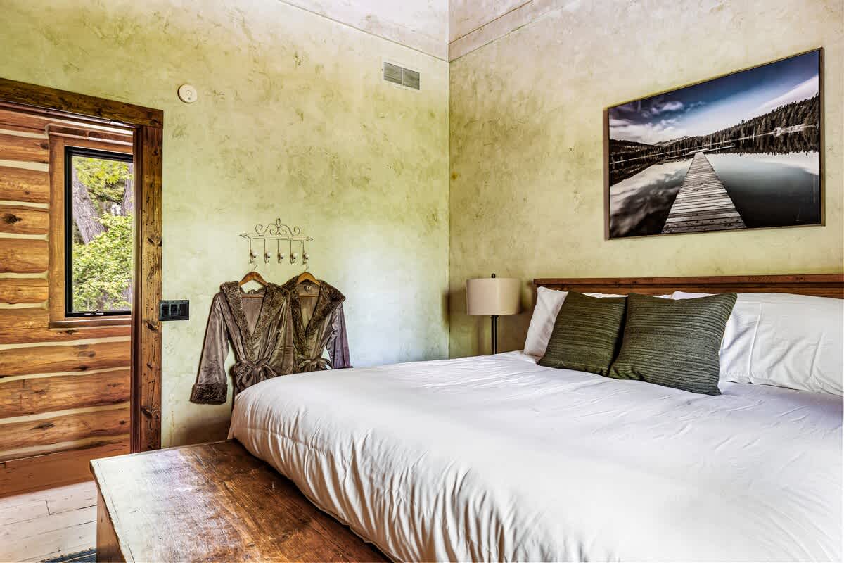 View of the cozy bedroom with a large bed, white bedding, and dark green pillows, showing a light green textured wall, rustic log walls, a wooden doorway, and two plush robes hanging on a wall rack.