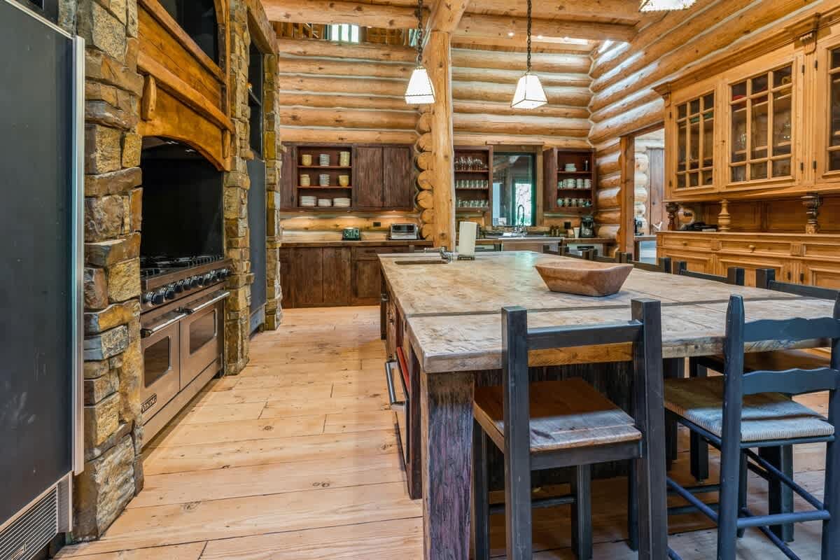 A closer view of the log cabin kitchen, highlighting the large wooden island with seating, a stainless steel range oven, and built-in shelving within the stone and wood cabinetry.