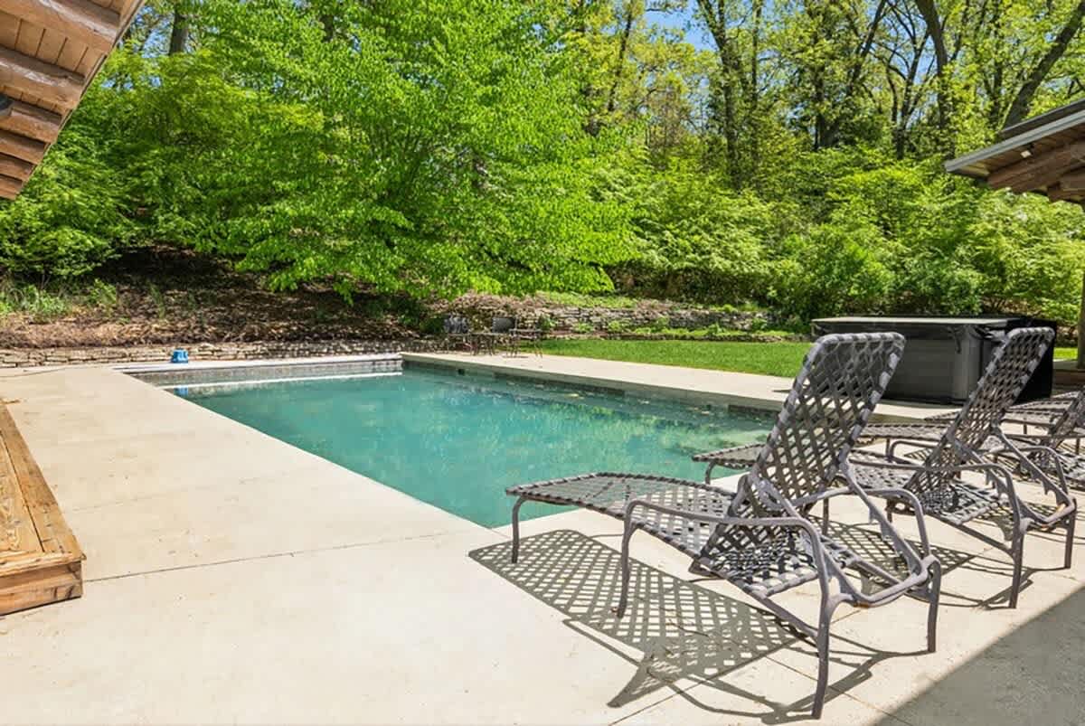 A rectangular outdoor swimming pool with clear blue water is surrounded by a spacious concrete patio, furnished with several dark mesh lounge chairs. A lush green lawn and dense forest provide a natural, private setting.