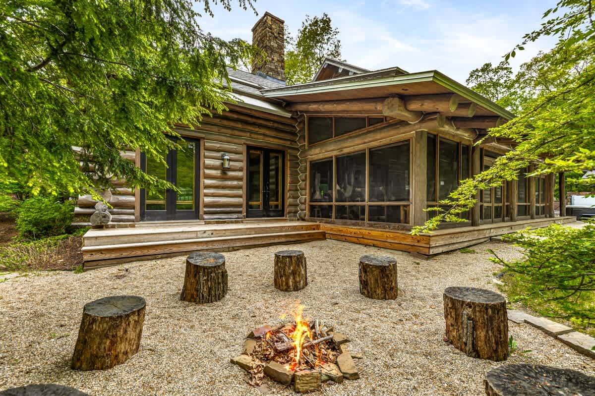 A cozy outdoor fire pit area with a crackling fire, surrounded by rustic tree stump seating on a gravel ground. The log cabin exterior with a screened porch is visible in the background, nestled among lush green trees.