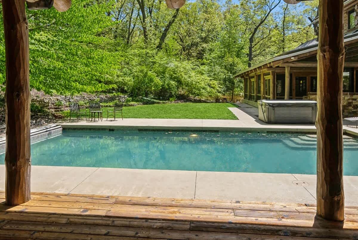A clear blue swimming pool is viewed from under a rustic wooden porch, with lounge chairs visible on the concrete patio. Lush green trees and foliage form a natural backdrop, creating a serene outdoor oasis.