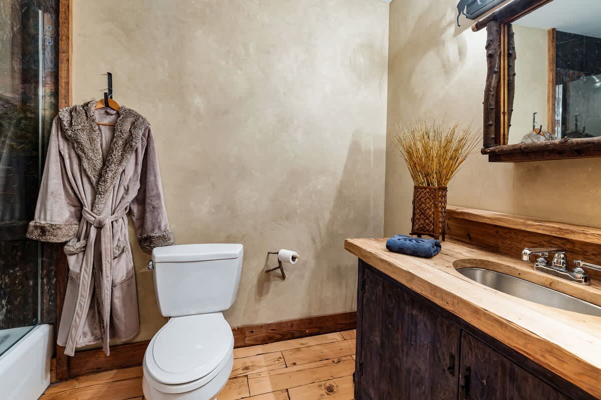Rustic bathroom with a single wooden vanity and sink, a log-framed mirror, a toilet, and a shower/tub combo, featuring textured beige walls and a plush robe hanging on the wall.