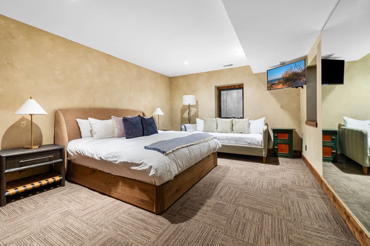 Modern bedroom with a large wooden-framed bed, white bedding, and blue pillows, a daybed, and a wall-mounted TV, set on patterned carpet with textured beige walls.