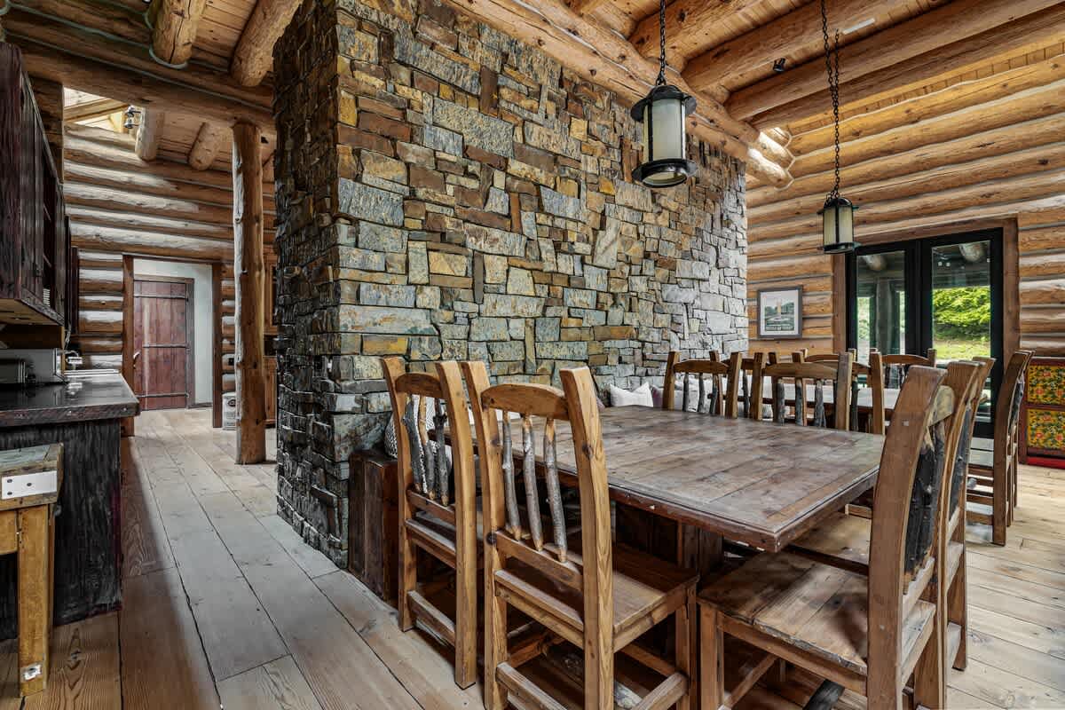 A rustic dining area with a large wooden table and matching chairs, set against a textured stone wall and surrounded by exposed log cabin architecture. Hanging lanterns provide ambient lighting.