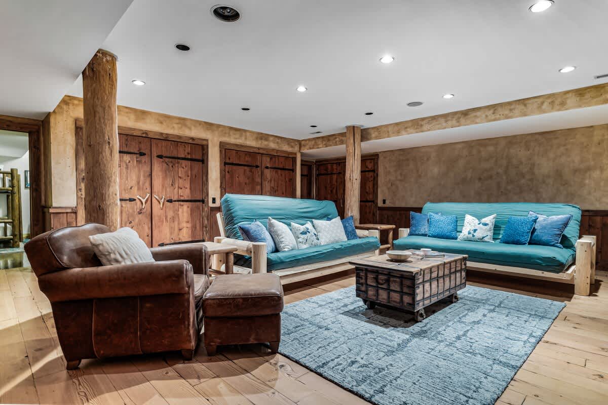 Cozy living room featuring two teal futons with decorative pillows, a leather armchair and ottoman, and a rustic wooden coffee table, all set on hardwood floors with a blue area rug.