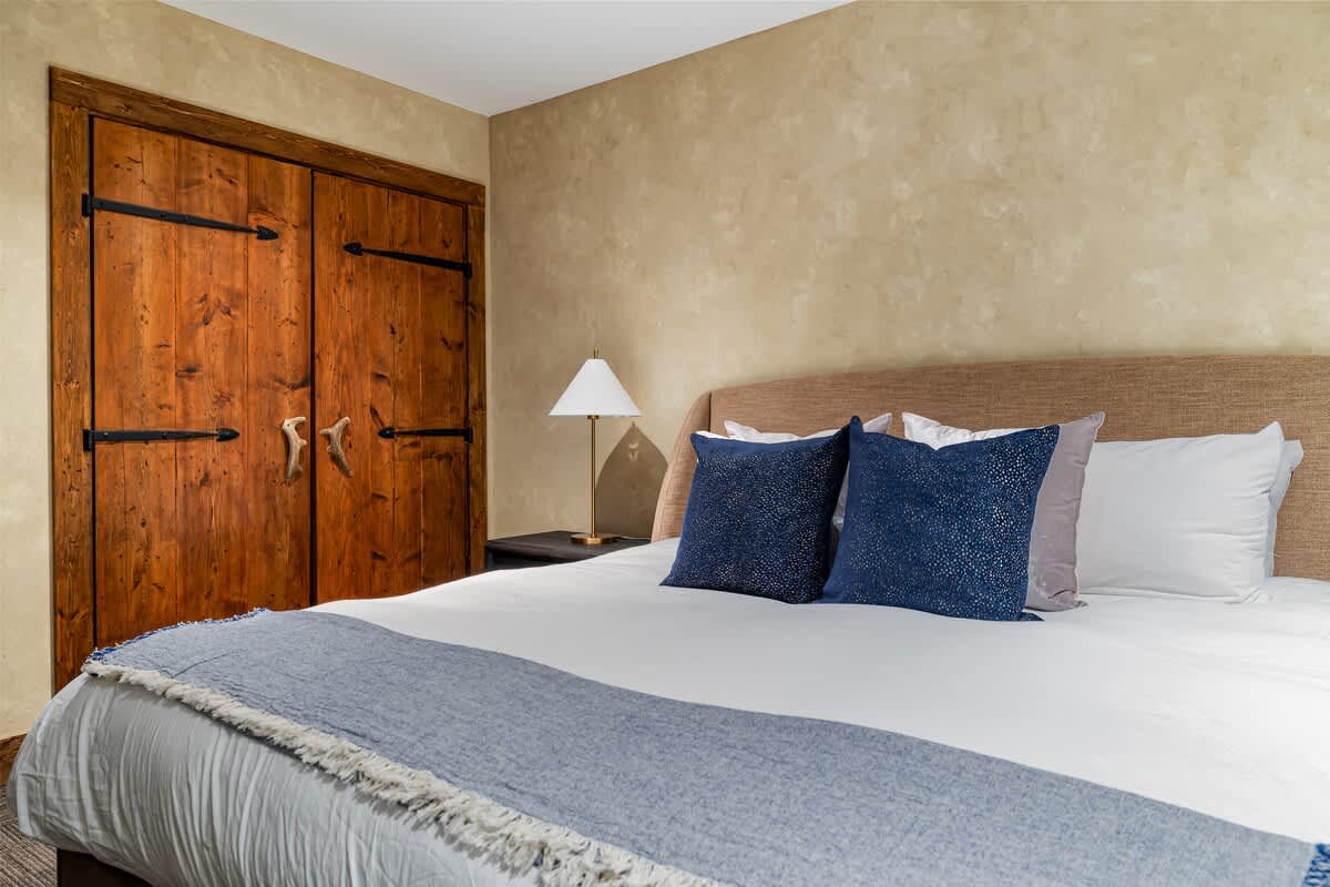 Close-up of the large bed with white bedding and blue pillows in a modern bedroom, featuring rustic wooden closet doors with black hardware against textured beige walls.