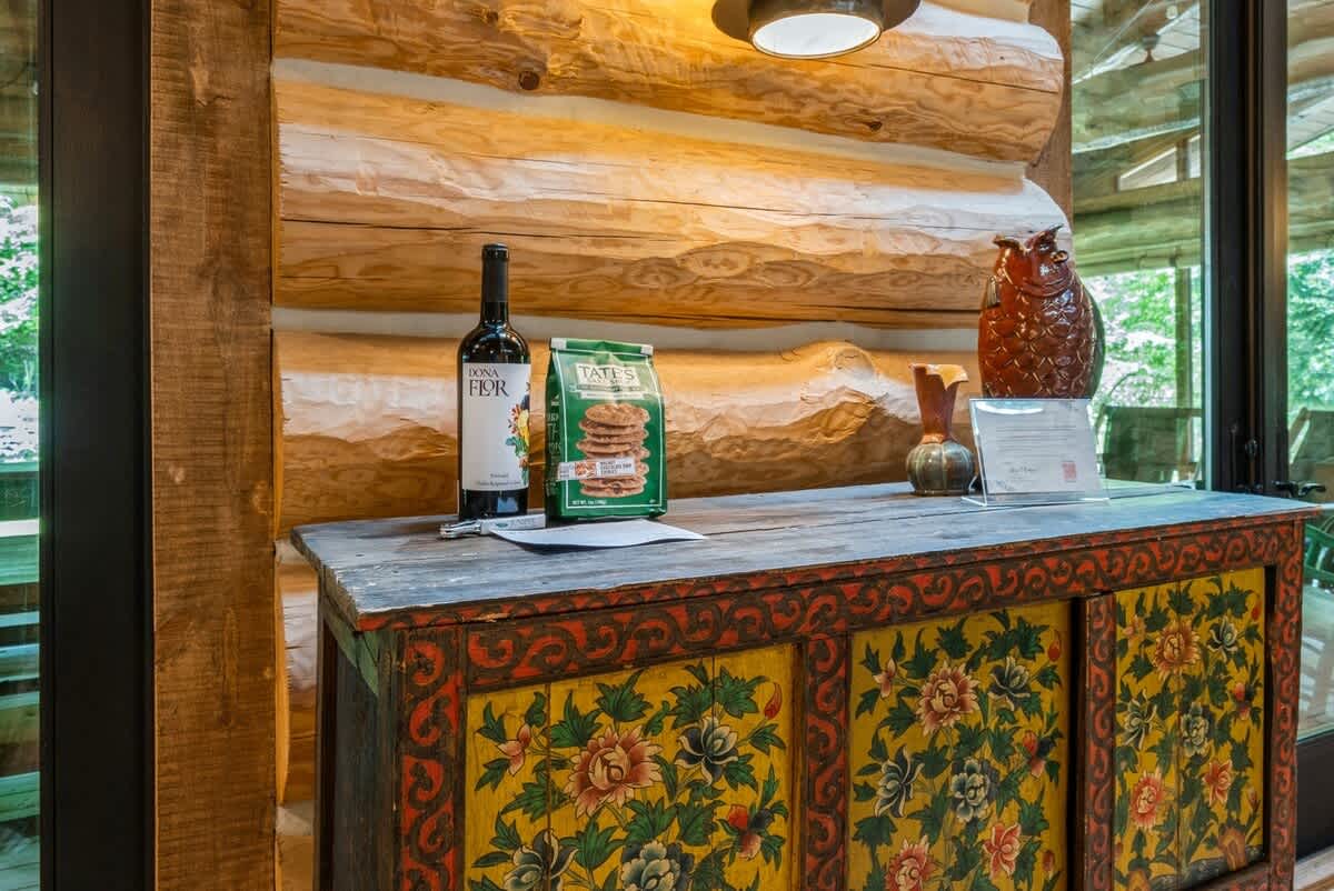 A welcoming console table with a rustic, hand-painted design, holding a bottle of wine, cookies, and decorative items. The background shows exposed log walls and a glimpse of an outdoor area, suggesting an entry or hallway space.