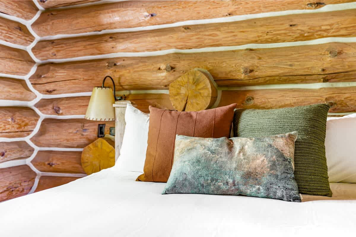 Close-up of a bed in a log cabin bedroom, showcasing decorative pillows in earthy tones against a white headboard and the textured log wall.
