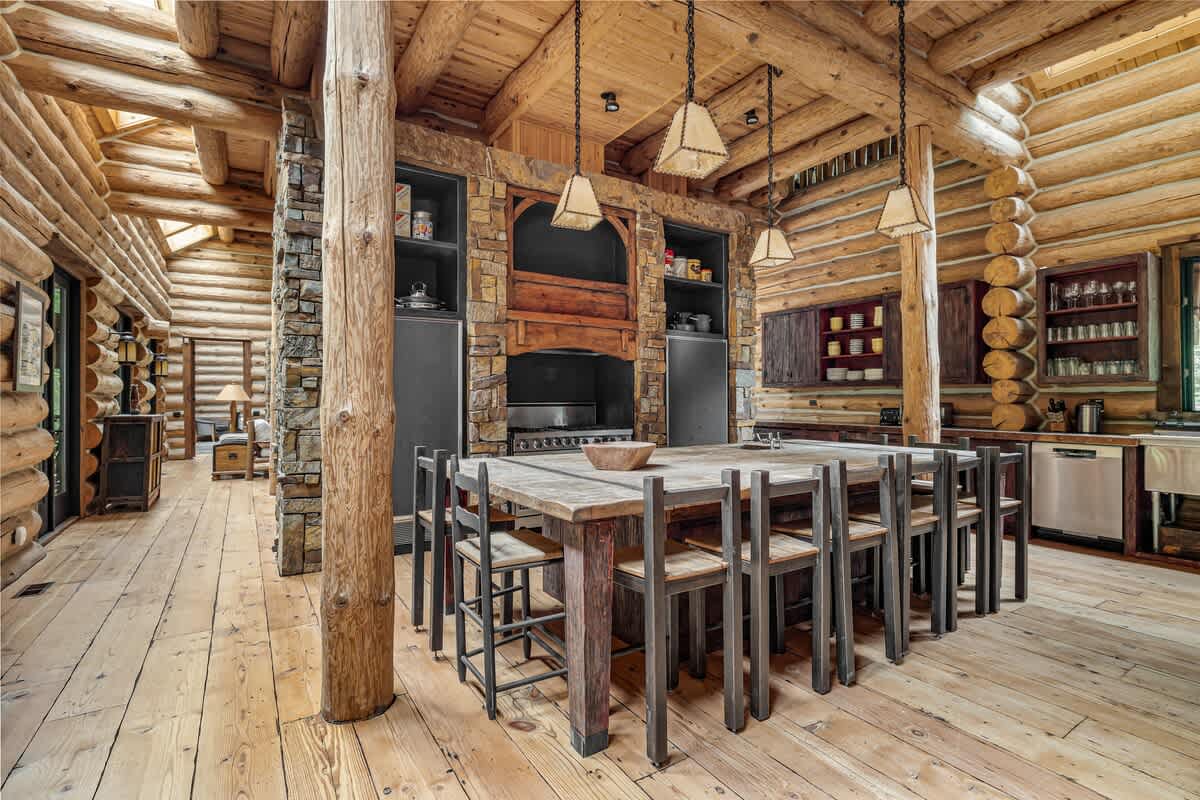 A spacious log cabin kitchen featuring a large wooden island with seating, a professional-grade range set into a stone alcove, and exposed log walls and ceiling.
