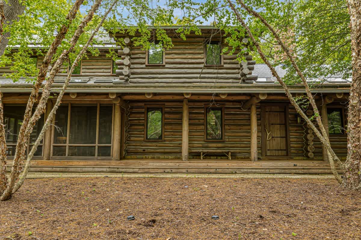 The front exterior of a charming rustic log cabin house, featuring a long wooden porch and multiple windows. The property is surrounded by mature trees and natural ground cover, highlighting its secluded and natural setting.