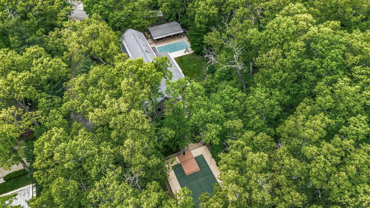 An aerial view showcases the property nestled within a dense green forest, featuring a main house with a swimming pool and a separate sports court, all surrounded by lush tree canopies.