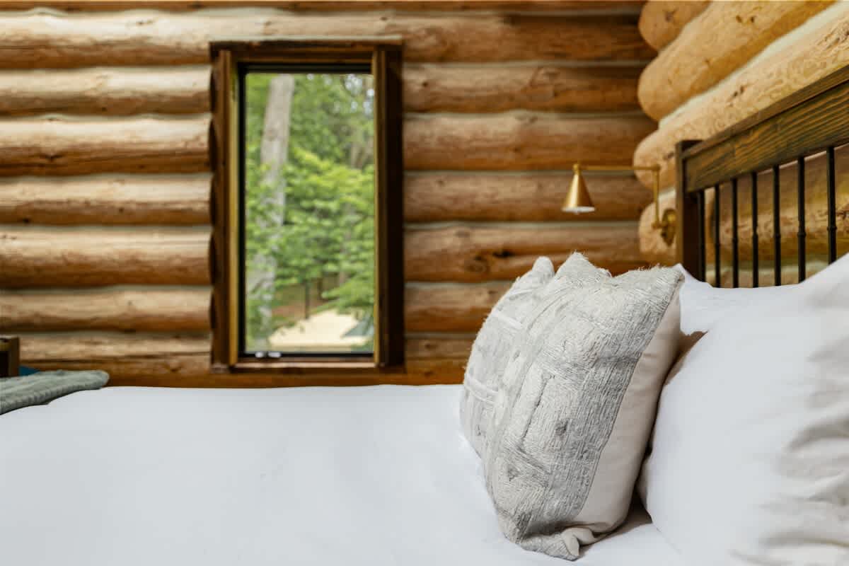 Close-up view of the comfortable bed in the log cabin bedroom, showcasing crisp white bedding, textured decorative pillows, and a narrow window offering a glimpse of the outdoors.