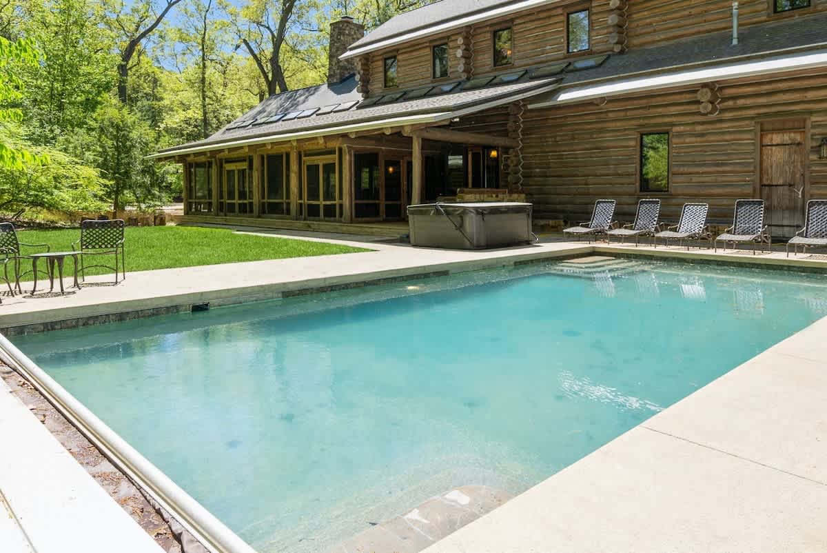 An inviting outdoor swimming pool with light blue water, surrounded by a concrete deck and lounge chairs, set against a rustic log cabin house with a hot tub nearby. Lush green trees frame the background, offering a serene natural setting.