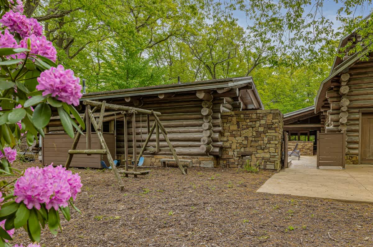 A rustic wooden swing set positioned on a mulched ground area, adjacent to a log cabin with stone accents, surrounded by blooming pink rhododendrons and lush trees.