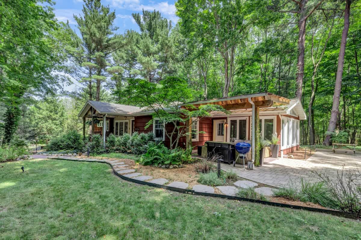The back of a rustic house with red siding is surrounded by tall trees and lush landscaping, featuring a paved patio with a grill and a covered outdoor seating area, accessible via a winding stone path through a green lawn.
