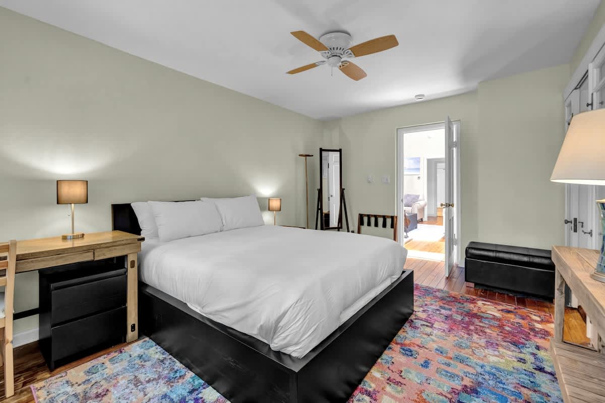 A cozy bedroom featuring a large black platform bed with white bedding, a vibrant multi-colored rug, and light green walls, with a ceiling fan and a desk area.