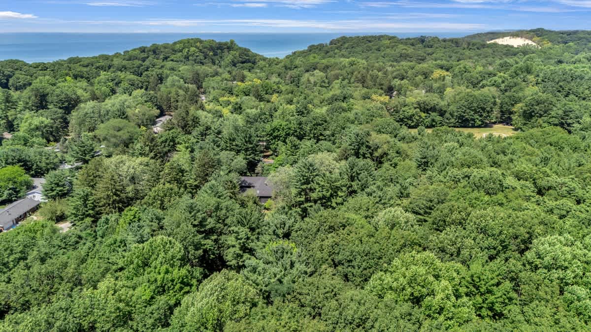 A wide aerial shot revealing the house surrounded by an expansive, lush green forest, with a distant view of a large body of water, likely Lake Michigan, and sand dunes under a clear sky.