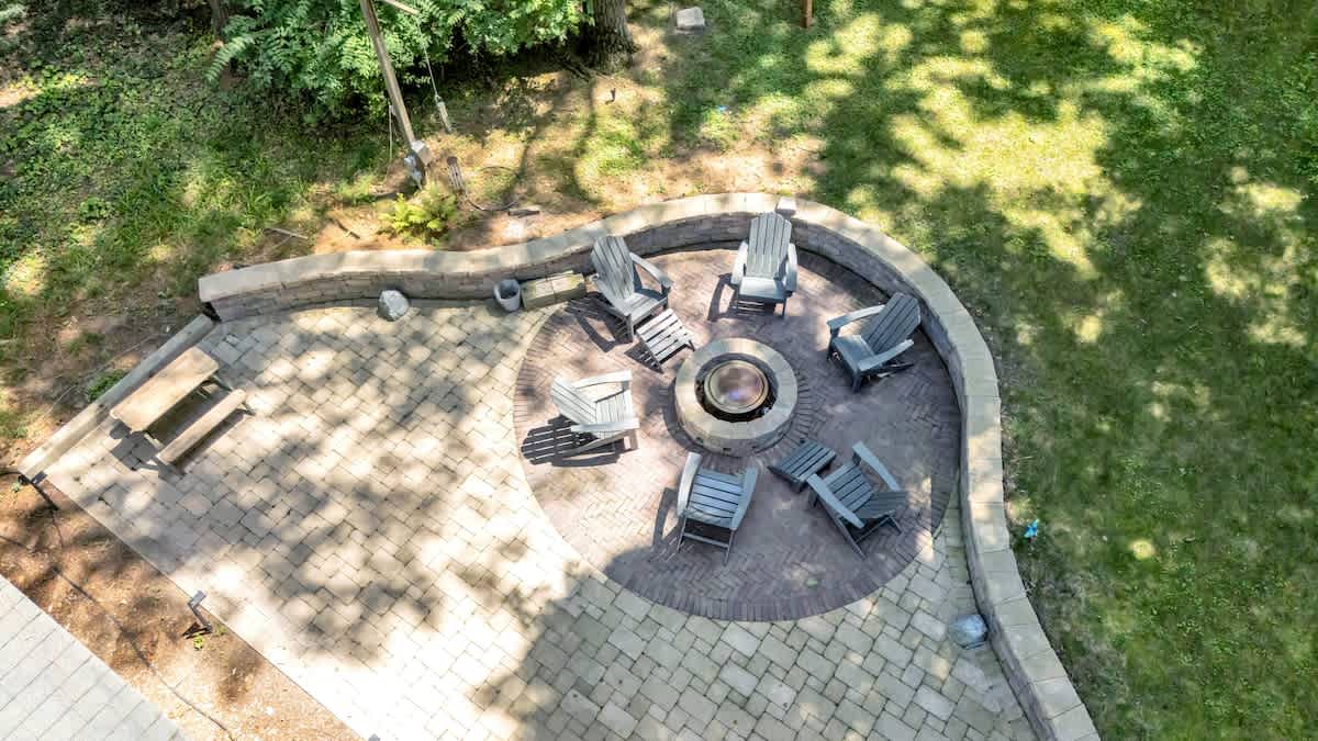 An overhead view of a large, curved brick paver patio featuring a central fire pit surrounded by Adirondack chairs. The patio is bordered by a low stone wall and set within a grassy yard.