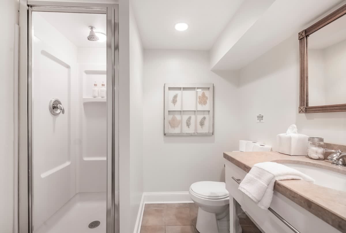 A bright white bathroom featuring a glass-enclosed shower with a white surround, a toilet, and a vanity with a light-colored countertop and a wooden-framed mirror. The floor is tiled in a light brown hue.