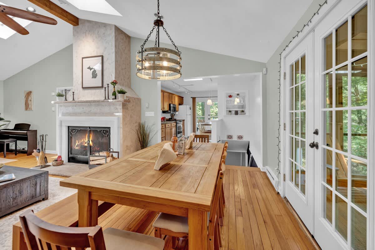 A bright dining area with a large wooden table and bench seating, featuring a modern chandelier and opening into a living room with a stone fireplace and vaulted ceiling.