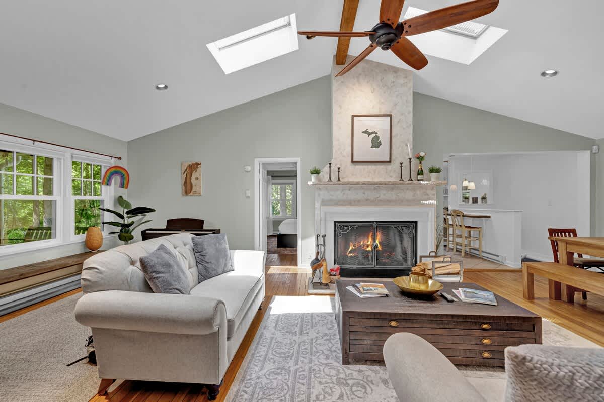 Spacious living room with a vaulted ceiling, skylights, and a cozy fireplace. A white tufted sofa sits on a patterned rug, with a piano and large windows offering views of greenery in the background.