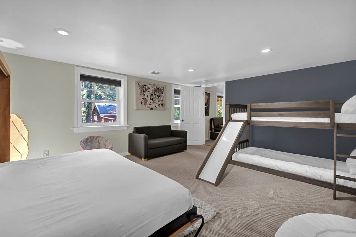 A spacious bedroom featuring a large bed with white linens, a wooden bunk bed with an attached slide, and a dark grey sofa, all set on light brown carpet. The room has light green and dark blue accent walls, with two windows offering views of trees.